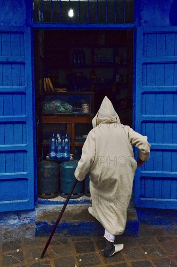 Old man with cane and hooded djellaba walks into shop