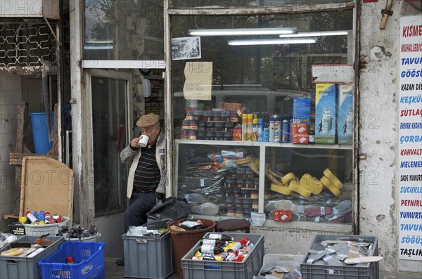 Old man drinking tea in front of his shop with consumer goods