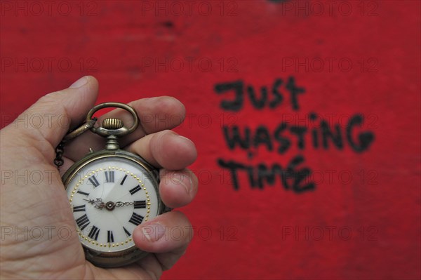 Hand with pocket watch in front of red wall with lettering just waisting time