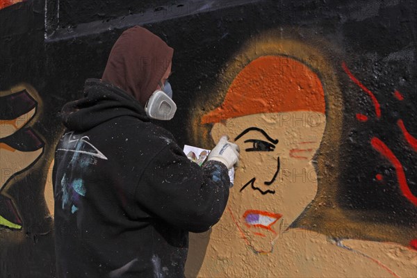 Graffiti sprayer dressed in black with hood and breathing protection in front of black wall with new work