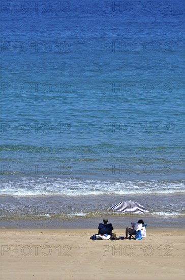 Man and woman under parasol on beach at water's edge from sea