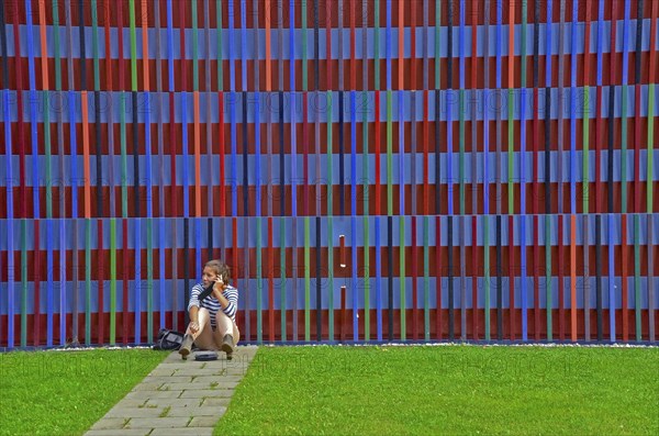 Young woman on the phone sitting on the floor in front of the colourful striped facade of the Museum Brandhorst