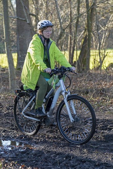 Woman cycling with e-bike over muddy forest path and through puddles