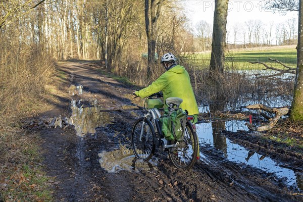 Woman cycling with e-bike over muddy forest path and through puddles