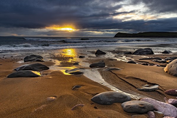 Sandy beach beach with stones on North Atlantic with rocky outcrop and cloudy sky at sunset