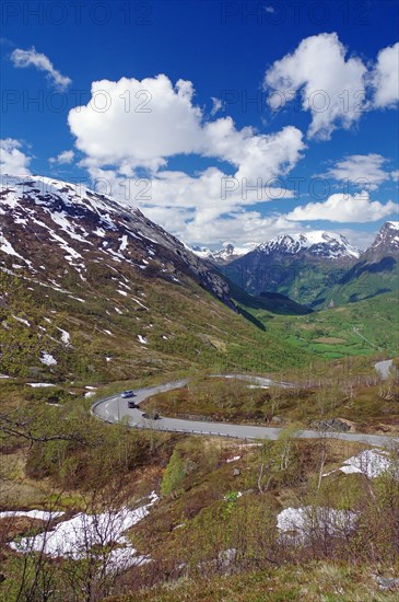 Road leads through spring-like landscape past partly snow-covered mountains downhill