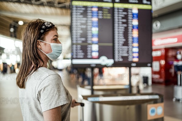 Ayoung girl in a mask waiting at the airport with flight information board as the background