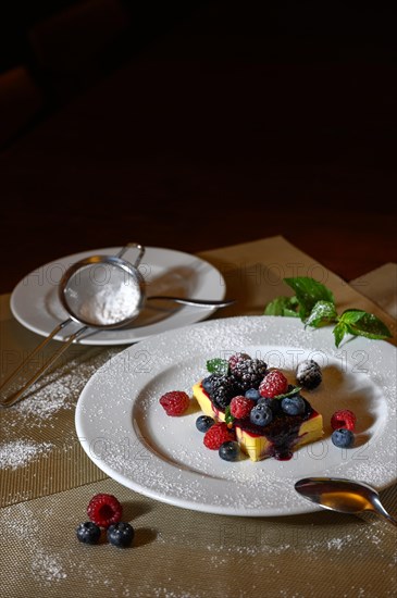 A piece of cheesecake with fresh raspberries and blackberries and a layer of icing sugar on a white plate. Next to it a sieve for applying the sugar