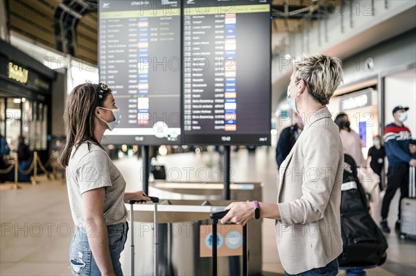 Two travelling women wearing protective masks discussing by flight information board at the Faro airport