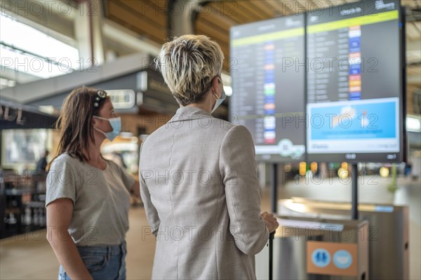 Two travelling women wearing protective masks discussing by flight information board at the Faro airport