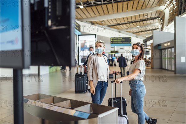 Two travelling women wearing protective masks discussing by flight information board at the Faro airport