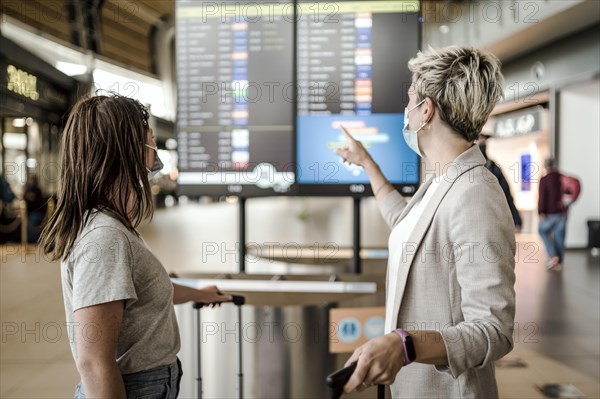 Two travelling women wearing protective masks discussing by flight information board at the Faro airport