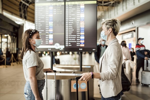 Two travelling women wearing protective masks discussing by flight information board at the Faro airport