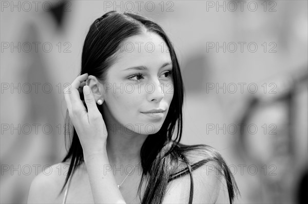 Black and white portrait of a very young pretty woman with a blurred background. With her right hand she guides her long hair behind her ear