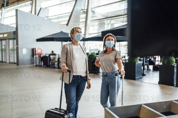 Two travelling women wearing protective masks discussing by flight information board at the Faro airport