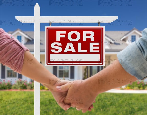 A couple hold his hands in front of a beautiful new house with sign for sale