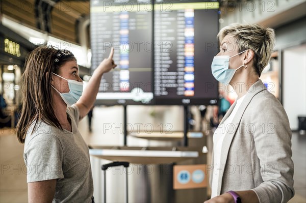 Two travelling women wearing protective masks discussing by flight information board at the Faro airport