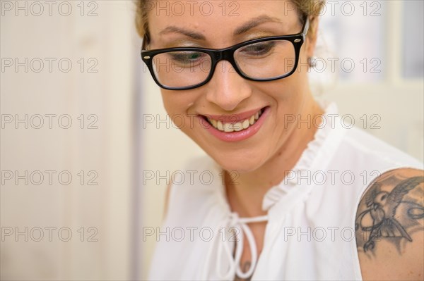 Portrait of a pretty young woman with blonde hair tied back and large black glasses looks down with a smile and beautiful teeth. The sleeveless white blouse reveals a tattooed shoulder