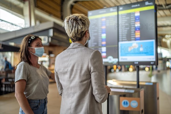 Two travelling women wearing protective masks discussing by flight information board at the Faro airport