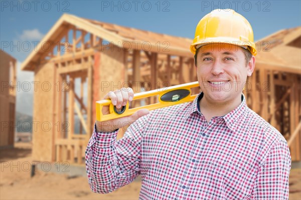 Smiling male contractor in hardhat holding blueprints and level at home construction site