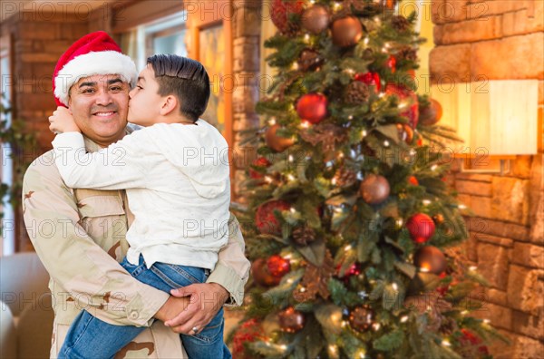 Hispanic armed forces soldier wearing santa hat hugging son in front christmas tree