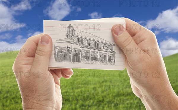 Male hands holding stack of paper with house drawing over empty grass field and sky