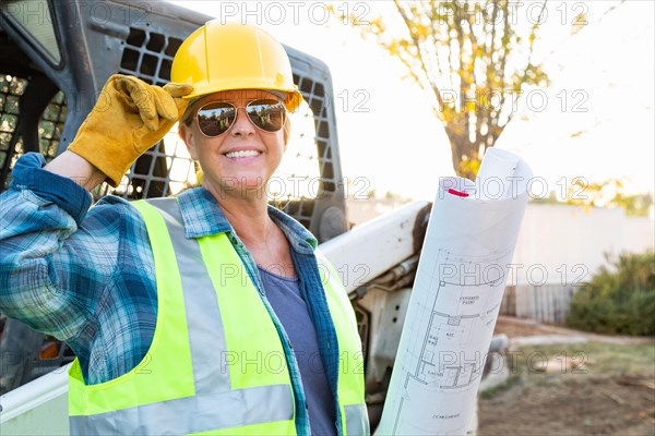 Smiling female worker holding technical blueprints near small bulldozer at construction site