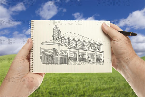 Male hands holding pen and pad of paper with house drawing over empty grass field and sky