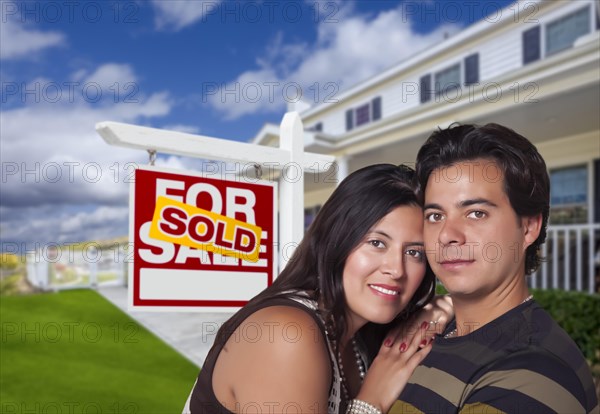 Young happy hispanic couple in front of their new home and sold for sale real estate sign