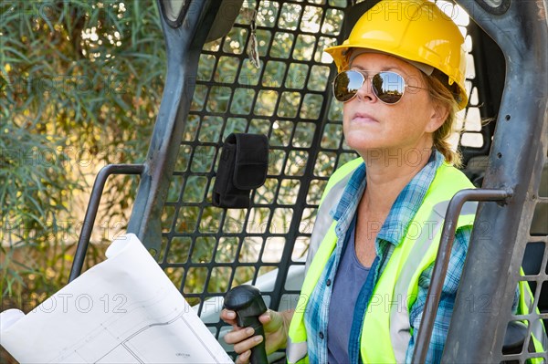 Smiling female worker holding technical blueprints using small bulldozer at construction site