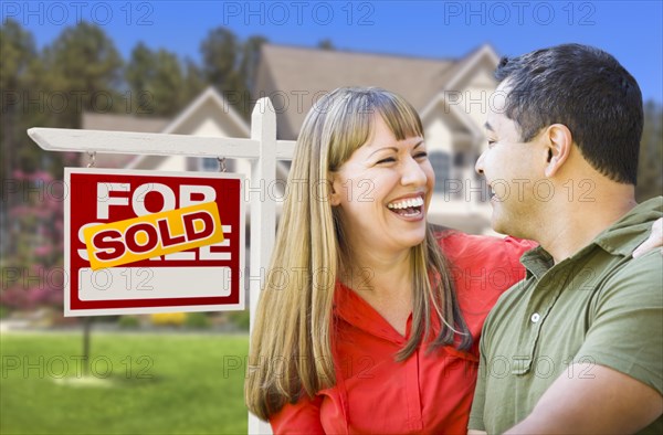 Happy mixed-race couple in front of sold home for sale real estate sign and house