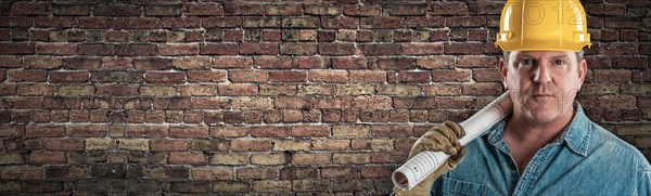 Male contractor in hard hat holding construction plans in front of old brick wall banner with copy space