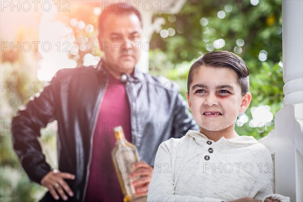 Afraid and bruised mixed-race boy in front of angry man holding bottle of alcohol