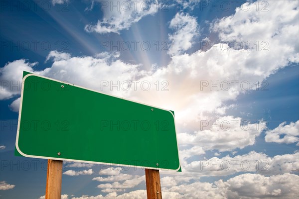 Blank green road sign over dramatic blue sky with clouds and sunburst