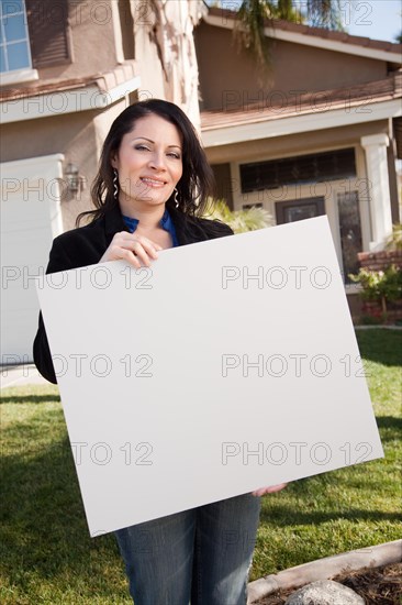 Happy attractive hispanic woman holding blank sign in front of house