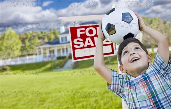 Playful young boy holding soccer ball in front of house and for sale real estate sign