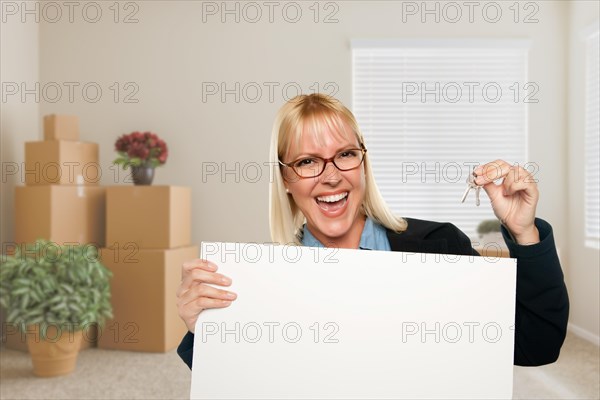 Happy attractive woman with blank sign and house key in empty room with packed moving boxes and potted plants