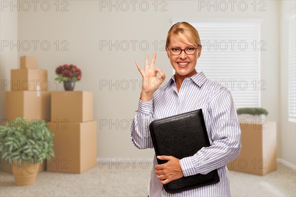 Female real estate agent with okay sign and binder in empty room with packed moving boxes and plants