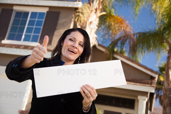 Happy attractive hispanic woman with thumbs up holding blank sign in front of house