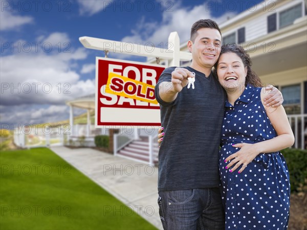 Happy hispanic couple in front of new home and sold real estate sign showing off their house keys