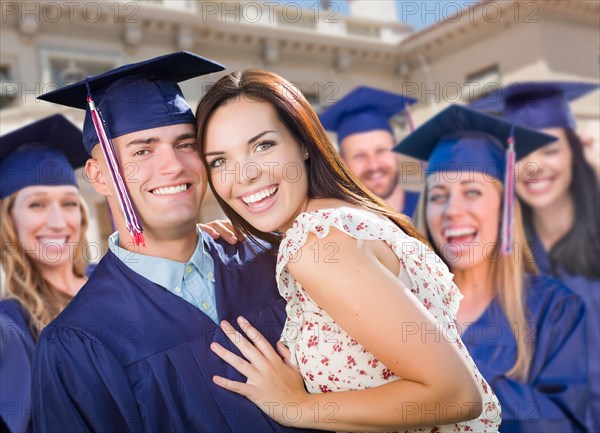 Proud male graduate in cap and gown with girl among other graduates behind