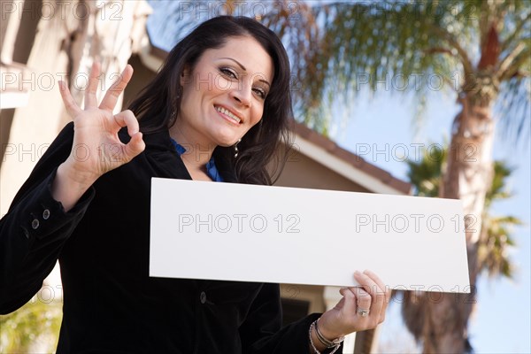 Happy attractive hispanic woman holding blank sign in front of house