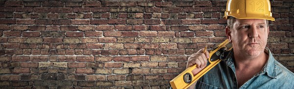 Male contractor in hard hat holding level in front of old brick wall banner with copy space