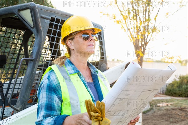 Female worker holding technical blueprints near small bulldozer at construction site