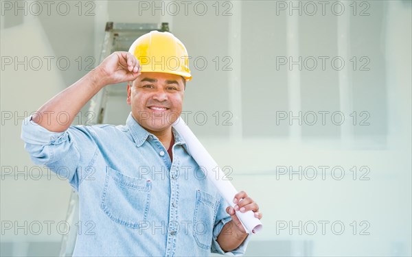 Hispanic male contractor with blueprint plans wearing hard hat in front of drywall and ladder