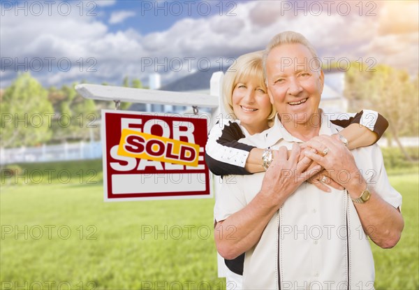 Happy affectionate senior couple hugging in front of sold real estate sign and house