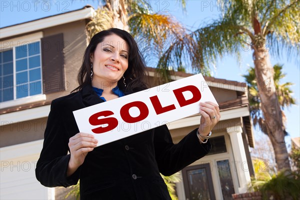 Happy attractive hispanic woman holding sold sign in front of house