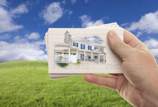 Male hand holding stack of paper with house drawing over empty grass field and sky