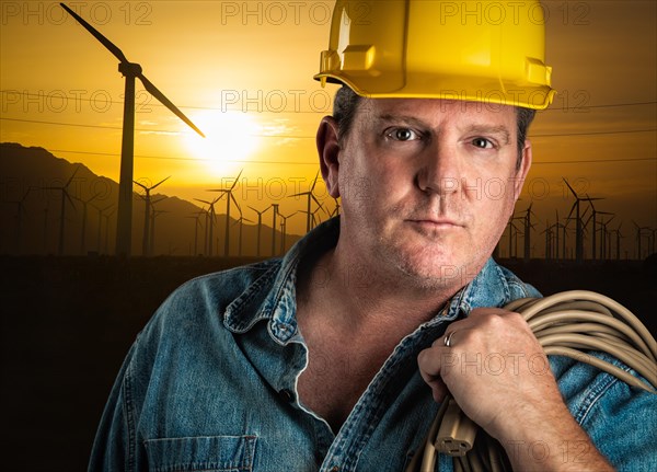 Serious contractor in hard hat holding extension cord outdoors near wind turbines