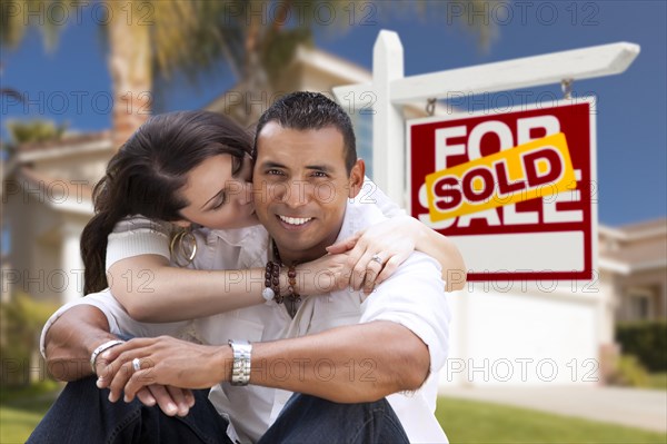 Young happy hispanic young couple in front of their new home and sold for sale real estate sign